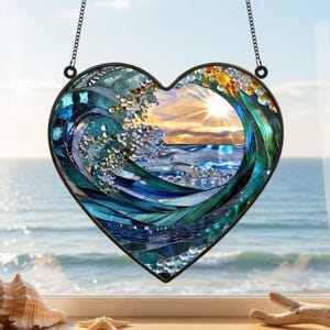 May include: A heart-shaped stained glass suncatcher featuring a detailed ocean wave scene with shades of blue, teal, and green glass pieces. The center depicts a sunset over the water with a bright sunburst, all framed by a dark metal border.