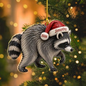Peut inclure: Une d&eacute;coration de sapin de No&euml;l en forme de raton laveur gris et noir portant un bonnet de P&egrave;re No&euml;l rouge &agrave; bordure blanche. Le bonnet est brod&eacute; du nom Jimothy et de l'ann&eacute;e 2026. La d&eacute;coration est suspendue par un cordon dor&eacute;.