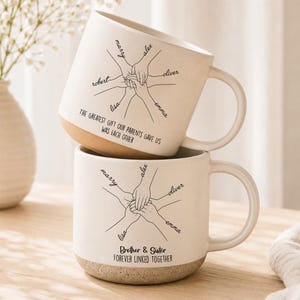 May include: Two off-white ceramic mugs with a speckled base. Each mug features a black line drawing of hands stacked together with names written along the arms. The top mug reads, "THE GREATEST GIFT OUR PARENTS GAVE US WAS EACH OTHER." The bottom reads, "Brother & Sister FOREVER LINKED TOGETHER."