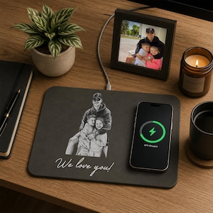May include: A rectangular black wireless charging pad featuring a custom white line-art illustration of a person holding two children, with the text "We love you!" printed below. A black smartphone is placed on the right side, showing a green charging icon.