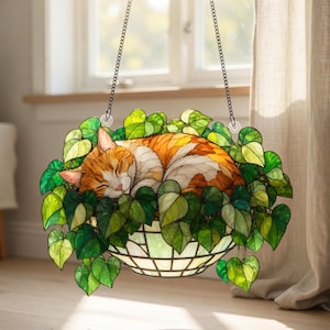 May include: A stained glass hanging planter featuring a sleeping orange and white tabby cat nestled among lush green heart-shaped leaves. The piece is suspended by two metal chains and includes a semi-circular glass bowl base with a grid pattern.