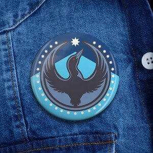 May include: A round button with a bird emblem in shades of gray and blue, set against a blue background. The bird is depicted with outstretched wings, a star, and mountain peaks. The button is on a denim jacket.