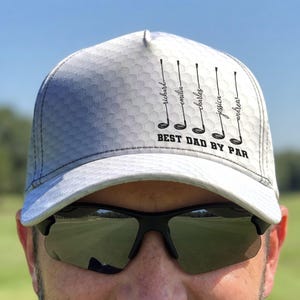 May include: A white baseball cap with a gray honeycomb pattern. The front features five black golf club illustrations, each labeled with the names Richard, Emilia, Charles, Jessica, and Andrew. Below the clubs, the text reads BEST DAD BY PAR.