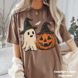 May include: A brown t-shirt with a Halloween design. The graphic features a ghost and a pumpkin, both wearing witch hats. Bats and stars are also included in the design. The text "Comfort Colors Espresso" is printed at the bottom of the shirt.