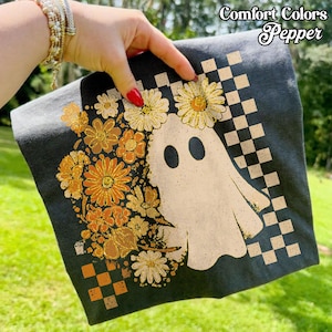 Peut inclure: T-shirt gris foncé avec un motif de fantôme crème orné d'une couronne florale et d'un motif à carreaux. Les fleurs sont orange et jaunes. Le texte "Comfort Colors Pepper" est en haut à droite.