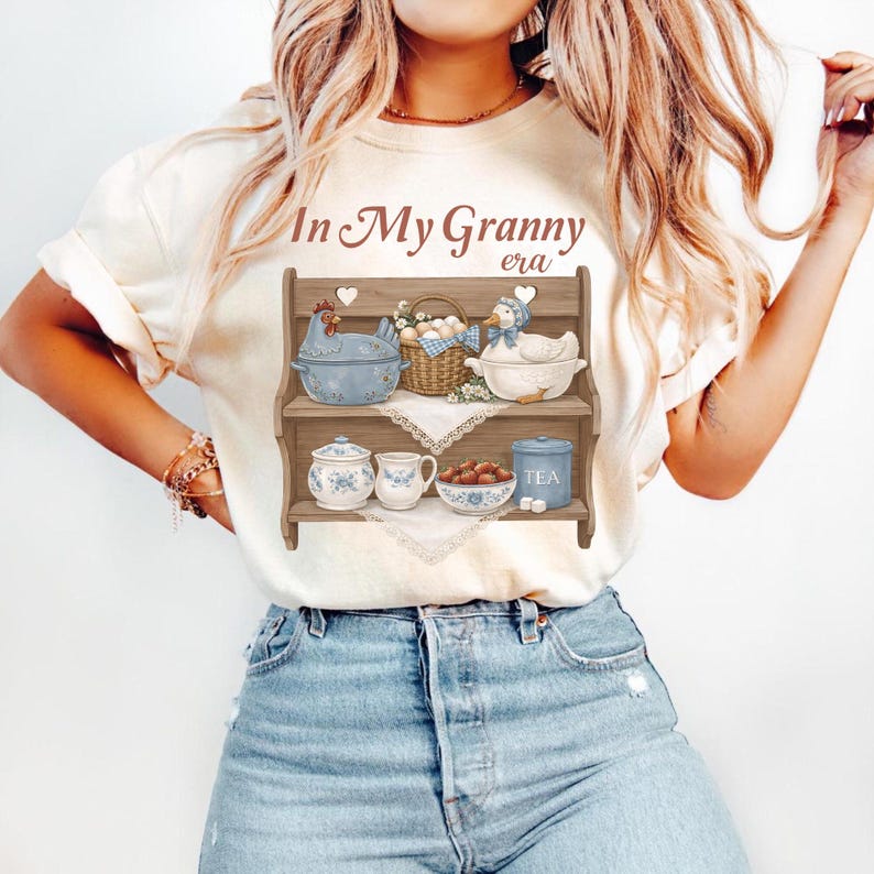 May include: Cream-colored t-shirt featuring a graphic of a wooden shelf with decorative items. The shelf holds a blue chicken, a basket of eggs, and a duck. Below are a sugar bowl, a teacup, a bowl of treats, and a tea canister. Text reads "In My Granny era."