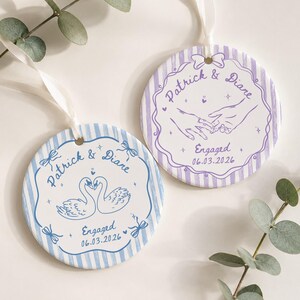 K&ouml;nnte beinhalten: Zwei runde Keramikornamente mit wei&szlig;en B&auml;ndern. Eines zeigt blaue Streifen, eine Schwanenillustration und den Text Patrick & Diare Engaged 06.03.2026. Das andere hat violette Streifen, eine Illustration von sich haltenden H&auml;nden und denselben Text.