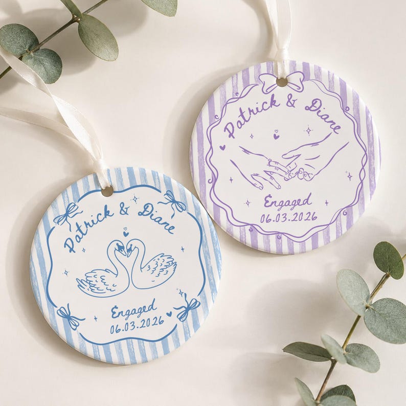 K&ouml;nnte beinhalten: Zwei runde Keramikornamente mit wei&szlig;en B&auml;ndern. Eines zeigt blaue Streifen, eine Schwanenillustration und den Text Patrick & Diare Engaged 06.03.2026. Das andere hat violette Streifen, eine Illustration von sich haltenden H&auml;nden und denselben Text.