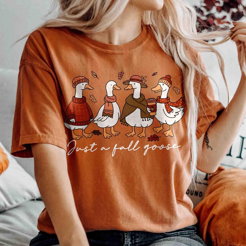 May include: Burnt orange t-shirt featuring a whimsical illustration of four geese dressed in fall attire. The geese are wearing hats, scarves, and holding a coffee cup. The text "Just a fall goose" is written in a cursive font below the geese.