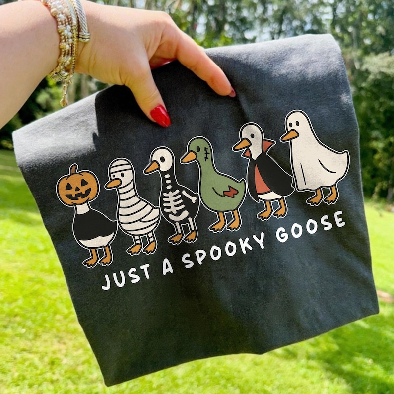 K&ouml;nnte beinhalten: Dunkelgraues T-Shirt mit einem Halloween-Motiv. Es zeigt Cartoon-G&auml;nse in Kost&uuml;men: K&uuml;rbis, Mumie, Skelett, Vampir und Geist. Der Text "JUST A SPOOKY GOOSE" ist darunter aufgedruckt.