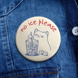 May include: A circular, cream-coloured button featuring a blue illustration of a disgruntled cat and a glass of ice cubes. The red text "no ice please" is written around the top of the button.
