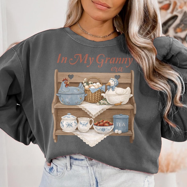 May include: Dark gray sweatshirt with a graphic of a wooden shelf displaying a blue chicken-shaped bowl, a basket of eggs, and a duck-shaped bowl. The text "In My Granny era" is printed above the shelf.