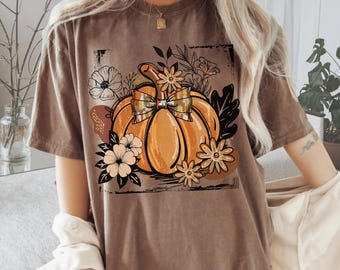 May include: Espresso-colored t-shirt featuring a pumpkin graphic with a plaid bow, surrounded by flowers and foliage. The text "Comfort Colors" and "Espresso" are printed at the bottom. A fall-themed graphic tee.