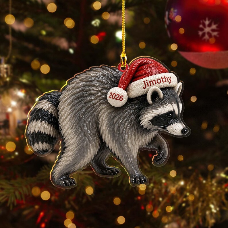 Peut inclure: Une d&eacute;coration suspendue plate en forme de raton laveur au pelage gris et noir, portant un bonnet de P&egrave;re No&euml;l rouge et blanc. Le bonnet porte le nom Jimothy en rouge et l'ann&eacute;e 2026 en rouge sur le pompon blanc. Suspendu par un cordon dor&eacute;.