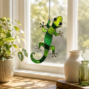 May include: A stained glass gecko suncatcher featuring various shades of green glass with a textured finish. The lizard is constructed with dark metal solder lines and includes a silver-toned metal chain for hanging from a window frame.