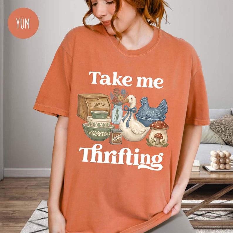 Op de afbeelding: Oranje T-shirt met korte mouwen met de tekst "Take me Thrifting" in het wit. De afbeelding bevat een brooddoos, kommen, een gans met een blauw lint, een blauwe kip en een paddenstoel.