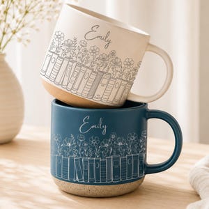 May include: Two ceramic mugs stacked vertically. The top mug is cream-colored with a gray line drawing of books and flowers, featuring the name Emily. The bottom mug is dark blue with a white line drawing of books and flowers, also labeled Emily.