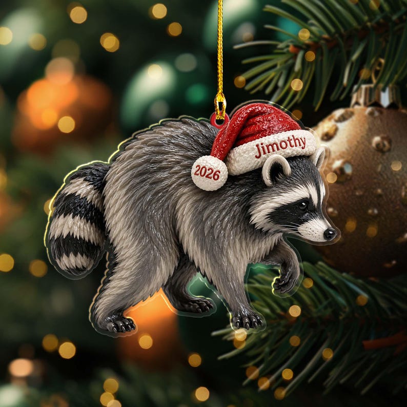 Peut inclure: Une d&eacute;coration en forme de raton laveur au pelage gris et noir, portant un bonnet de P&egrave;re No&euml;l rouge et blanc. Le bonnet est brod&eacute; du nom Jimothy et de l'ann&eacute;e 2026. La d&eacute;coration est suspendue par un cordon dor&eacute; et repr&eacute;sente l'animal en marche.