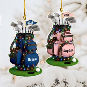 May include: Two hanging ornaments shaped like golf bags, one blue and one pink, each decorated with colourful string lights. The blue bag features the name Brian and the year 2025, while the pink bag features the name Sophia and the year 2025.