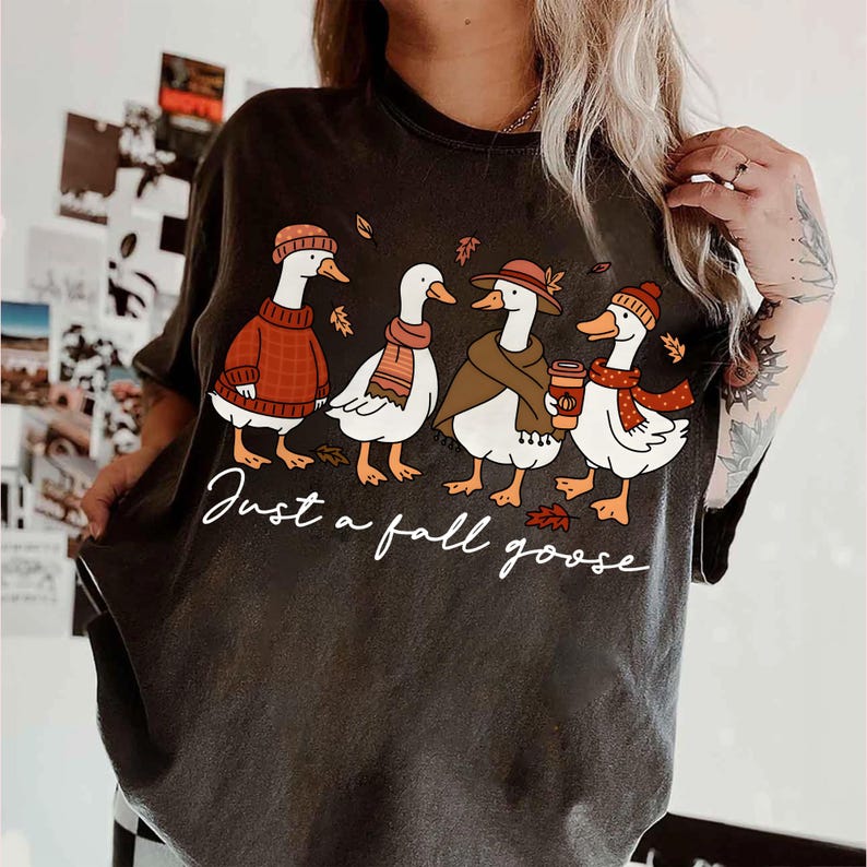 May include: Dark gray t-shirt featuring a whimsical illustration of four geese dressed in fall attire. The geese are wearing sweaters, scarves, and hats, with the text "Just a fall goose" in a cursive font.