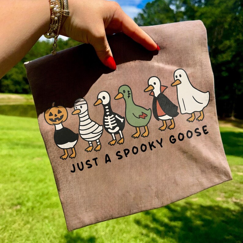 K&ouml;nnte beinhalten: Eine braune Canvas-Tragetasche mit einem Halloween-Design. Das Design zeigt sechs Cartoon-G&auml;nse in Kost&uuml;men, darunter ein K&uuml;rbis, eine Mumie, ein Skelett und ein Vampir. Der Text "JUST A SPOOKY GOOSE" ist darunter gedruckt.