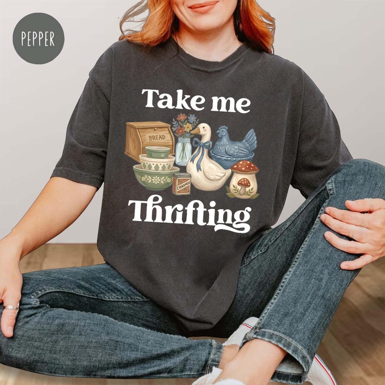 Op de afbeelding: Donkergrijs T-shirt met de tekst "Take me Thrifting" en een afbeelding van vintage keukenartikelen. De afbeelding bevat een brooddoos, kommen, een eend, een kip en een paddenstoel. Het shirt is gemaakt van zacht materiaal.