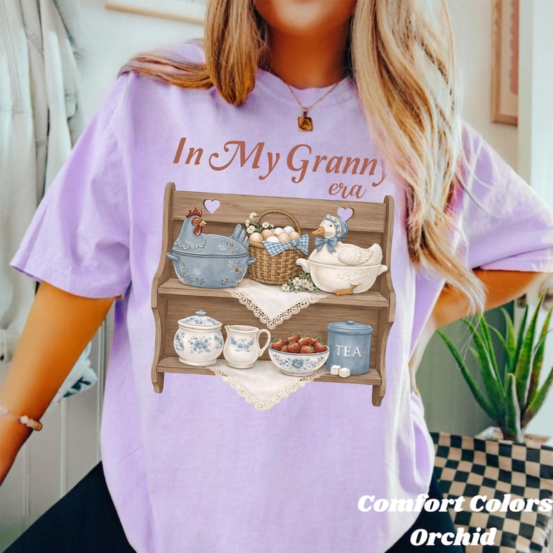 May include: A light purple t-shirt with the text "In My Granny era" and a graphic of a wooden shelf with kitchenware. The shelf features a chicken-shaped pot, a duck-shaped bowl, and a basket of eggs. The text "Comfort Colors Orchid" is also visible.