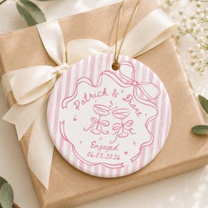 K&ouml;nnte beinhalten: Ein rundes, wei&szlig;es Keramikornament mit einem rosa gestreiften Rand und einer zentralen Illustration von zwei ansto&szlig;enden Sektgl&auml;sern mit Schleifen. Der Text lautet Patrick & Diane, Engaged, sowie das Datum 06.03.2026 in rosa Schrift.