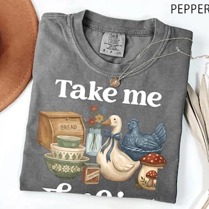 Op de afbeelding: Comfort Colors t-shirt in peperkleur met de witte woorden "Take me". De afbeelding toont een brooddoos, kommen, een eend met een blauwe strik, een blauwe kip, een paddenstoel en het woord "Cinnamon". Een zilveren ketting ligt over het shirt.