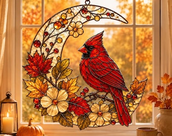May include: A crescent moon-shaped stained glass suncatcher featuring a vibrant red cardinal perched on a branch. The design includes autumn-colored maple leaves, white flowers, and small red berries, all framed in dark metal leading.