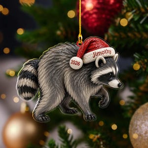 Peut inclure: Une d&eacute;coration de No&euml;l en forme de raton laveur avec une queue ray&eacute;e grise et noire, portant un bonnet de P&egrave;re No&euml;l rouge avec un pompon blanc. Le bonnet porte l'inscription Jimothy et l'ann&eacute;e 2026 en rouge. Un cordon dor&eacute; est fix&eacute; au sommet.