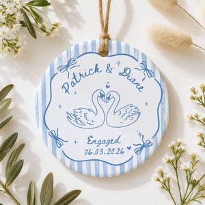 K&ouml;nnte beinhalten: Ein rundes Keramikornament mit hellblau-wei&szlig;em Streifenmuster. In der Mitte sind zwei wei&szlig;e Schw&auml;ne abgebildet, die sich ansehen, sowie der Text "Patrick & Diane", "Engaged" und das Datum "06.03.2026" in Blau mit dekorativen Schleifen.