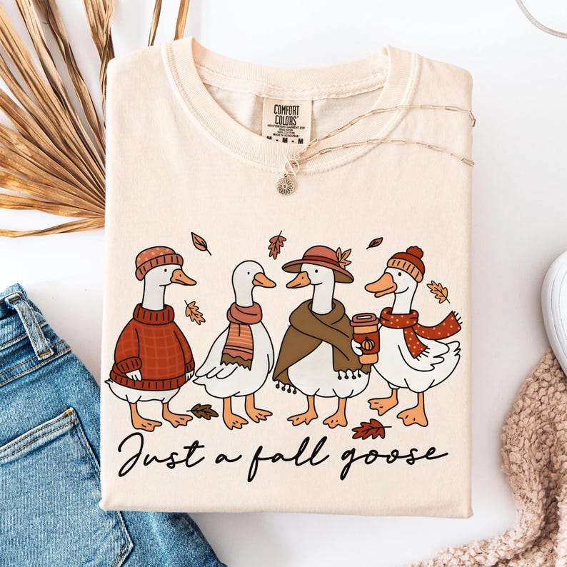 May include: Cream-colored t-shirt featuring a whimsical illustration of four geese dressed in fall attire. The geese wear hats, scarves, and sweaters, with one holding a coffee cup. The text "Just a fall goose" is written below the geese.