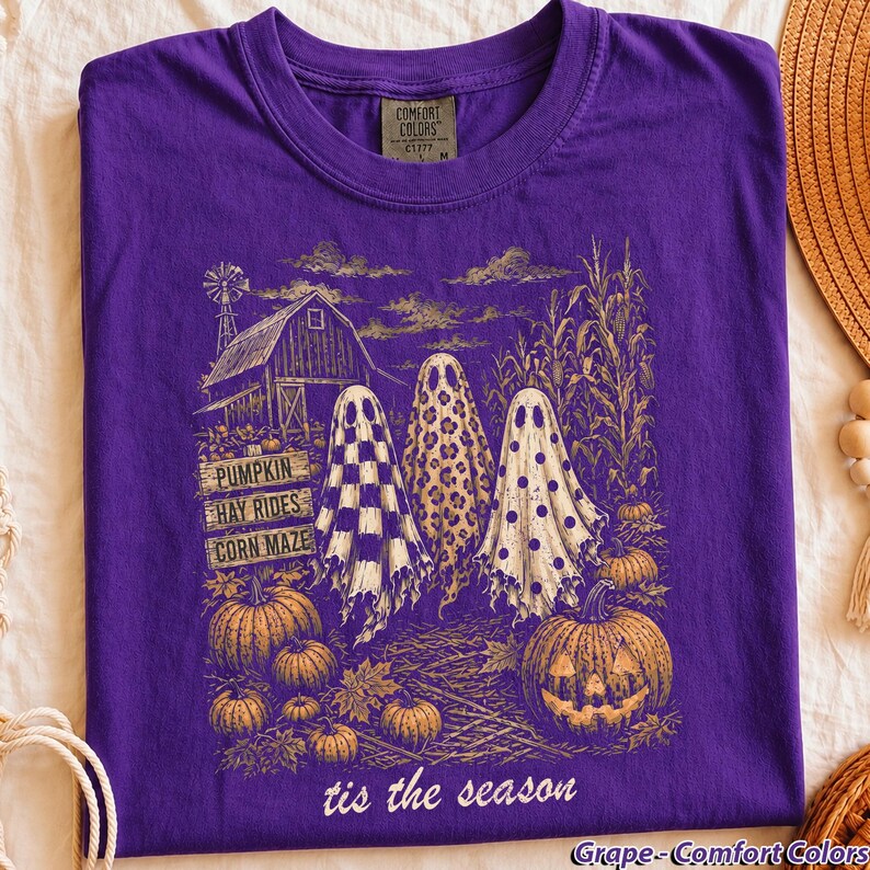 Pode incluir: Uma t-shirt roxa com uma estampa gr&aacute;fica de tr&ecirc;s fantasmas num campo com um celeiro, talos de milho e ab&oacute;boras. Os fantasmas t&ecirc;m padr&otilde;es xadrez, leopardo e bolinhas. O texto diz PUMPKIN, HAY RIDES, CORN MAZE e tis the season.