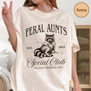 May include: Ivory-colored t-shirt with the text "FERAL AUNTS" above a raccoon illustration. Below the illustration, the text reads "Social Club" and "Exclusive Members Only". The shirt also has the text "EST. 2025".