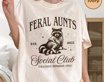 May include: Ivory-colored t-shirt with the text "FERAL AUNTS" above a raccoon illustration. Below the illustration, the text reads "Social Club" and "Exclusive Members Only". The shirt also has the text "EST. 2025".