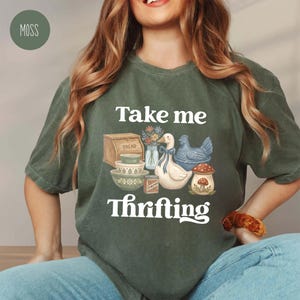Op de afbeelding: Mosgroen t-shirt met de witte tekst "Take me Thrifting". Het shirt heeft een afbeelding van een eend, kip, brooddoos, kommen en een paddenstoel. Het shirt is gemaakt van een zacht materiaal.