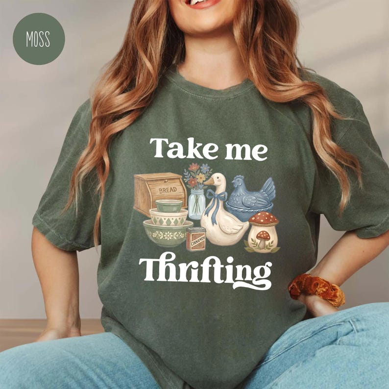 Op de afbeelding: Mosgroen t-shirt met de witte tekst "Take me Thrifting". Het shirt heeft een afbeelding van een eend, kip, brooddoos, kommen en een paddenstoel. Het shirt is gemaakt van een zacht materiaal.