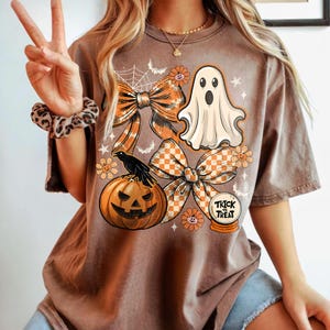 May include: Brown t-shirt featuring a Halloween design. The graphic includes orange bows, a ghost, a jack-o'-lantern, a crow, and the words "Trick or Treat" in a snow globe. The shirt has short sleeves.