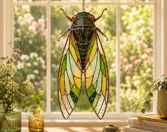 May include: A stained glass cicada suncatcher featuring a segmented body in shades of dark green and black with two prominent orange eyes. The wings are composed of translucent glass panels in varying hues of yellow, lime green, and clear glass.
