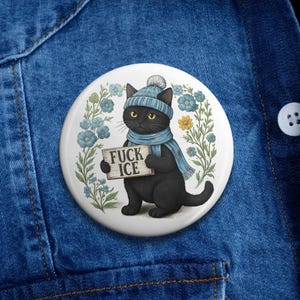 May include: A round white button featuring a black cat wearing a blue hat and scarf, holding a sign that reads "FUCK ICE." The cat is surrounded by blue and yellow floral accents.