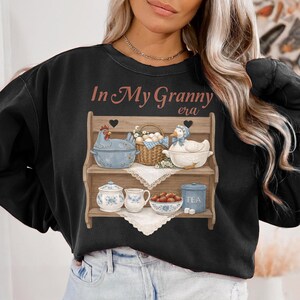 May include: Black sweatshirt with a graphic of a wooden shelf displaying kitchenware and the text "In My Granny era". The shelf holds a blue chicken-shaped bowl, a basket of eggs, and a duck-shaped bowl.