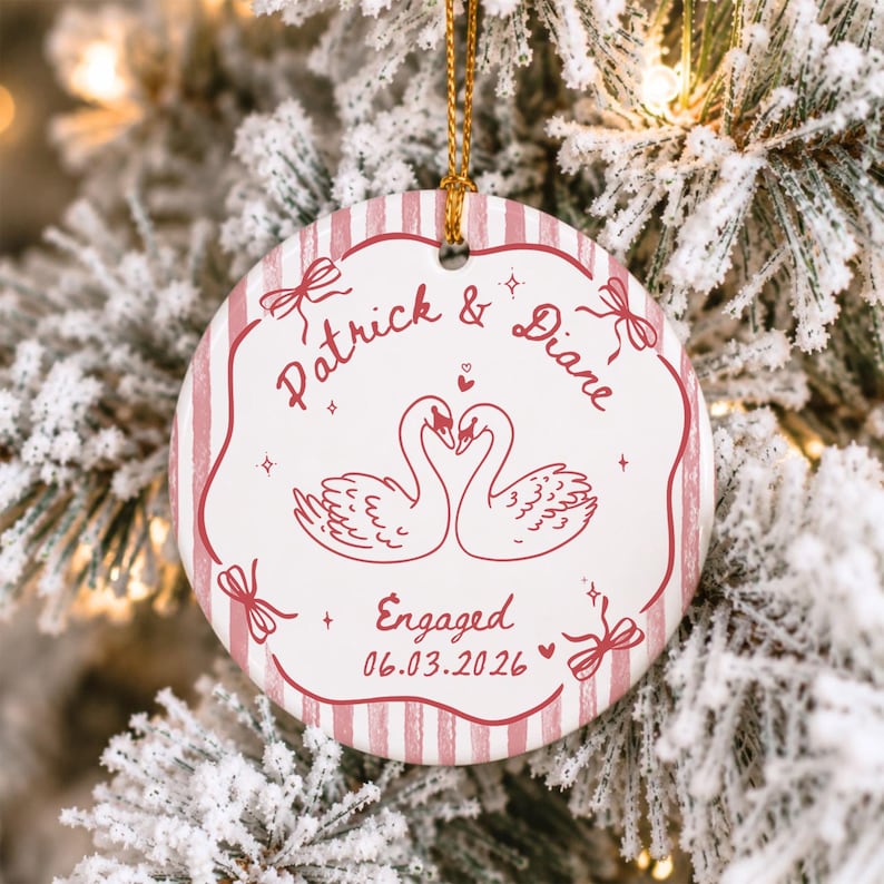 K&ouml;nnte beinhalten: Ein runder wei&szlig;er Keramikschmuck mit goldener Aufh&auml;ngeschnur, verziert mit einem rosa gestreiften Rand und einer zentralen Abbildung zweier Schw&auml;ne, die sich gegen&uuml;berstehen. Der Text lautet Patrick & Diane, Engaged, und das Datum 06.03.2026 in rosa Schrift.