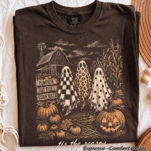 Pode incluir: Uma t-shirt castanho-escura com uma impress&atilde;o gr&aacute;fica de tr&ecirc;s fantasmas em frente a um celeiro e um milharal. Os fantasmas t&ecirc;m padr&otilde;es axadrezados, de leopardo e de bolinhas. O texto diz PUMPKIN, HAY RIDES, CORN MAZE e tis the season.