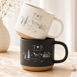 May include: A stacked pair of ceramic mugs, one cream and one black, each featuring a natural tan base. Both mugs are engraved with a mountain landscape, pine trees, a winding path, the initials T and S separated by a heart, and the date 10. 06. 2026.