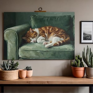 May include: A painting of a sleeping orange and white tabby cat on a green armchair. The artwork is displayed on a wall above a wooden table with potted plants. A framed black and white landscape photo hangs nearby.