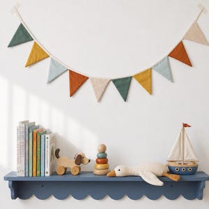 May include: A fabric bunting banner with triangular flags in muted shades of green, mustard yellow, light blue, rust orange, and beige, suspended above a blue scalloped shelf holding books, a wooden dog toy, a stacking ring toy, and a toy sailboat.