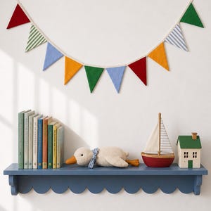 May include: A decorative bunting banner featuring triangular flags in red, green, blue, yellow, and striped patterns, suspended above a blue scalloped shelf holding a row of books, a plush duck with a blue gingham bow, a toy sailboat, and a small house.