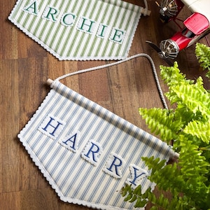 Op de afbeelding: Twee stoffen wandwimpels met witte touwophanging. Eén wimpel heeft groene verticale strepen met de naam ARCHIE in groene letters. De andere heeft blauwe verticale strepen met de naam HARRY in blauwe letters. Beide hebben geschulpte randen.