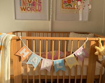 May include: A wooden baby crib with a banner that spells out "HARLOW" in pastel colors. The crib is in a nursery with framed art on the wall and a hanging lamp. A yellow blanket and a stuffed sun are also visible.